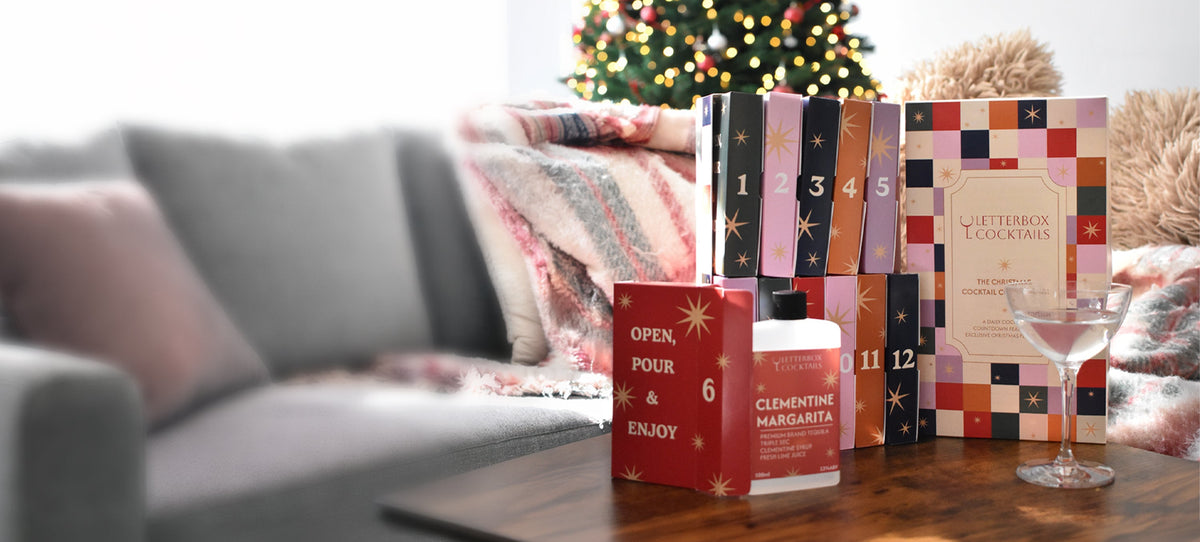 A Letterbox Cocktail Advent Calendar boxes arranged on a wooden table in a cozy living room setting with a blurred gray sofa, throw blanket, and a decorated Christmas tree in the background.