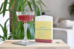 A bottle of 'Blood Orange Martini' next to a coupe glass filled with the deep red cocktail, set on a wooden table in a softly blurred living room with a green plant in the background.
