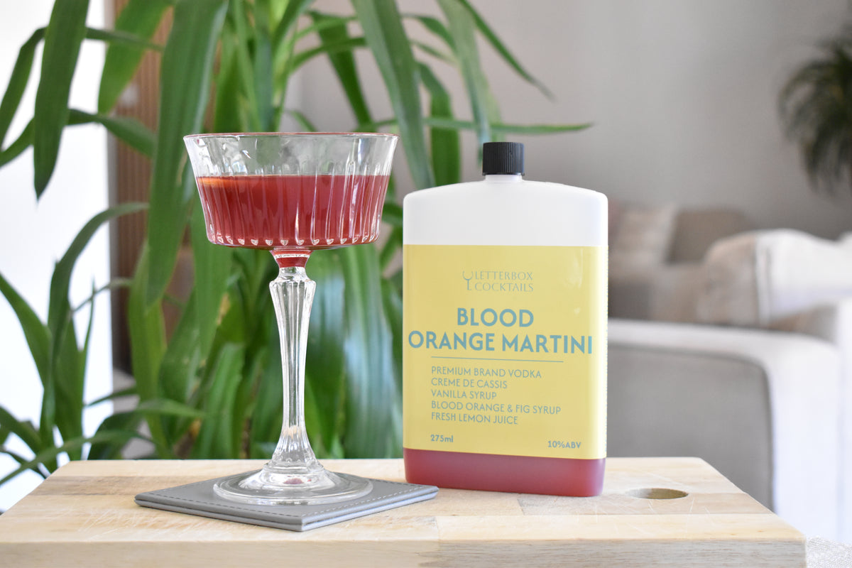 A bottle of 'Blood Orange Martini' next to a coupe glass filled with the deep red cocktail, set on a wooden table in a softly blurred living room with a green plant in the background.
