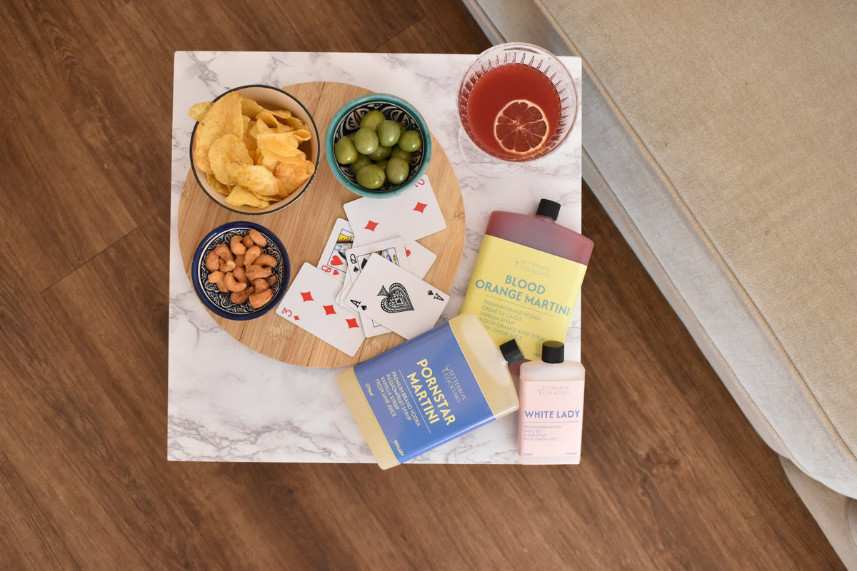 A styled flat-lay of a cocktail kit and snacks arranged on a white marble tray over a wooden table. The kit includes a Blood Orange Martini mix bottle, a White Lady cocktail , a Pornstar Martini cocktail, playing cards, olives, peanuts, chips, and a pink drink in a glass.