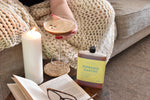 A cozy lifestyle scene showing a bottle of  espresso martini' on a wooden table next to a lit white pillar candle, an open book, and a person holding a martini glass wrapped in a chunky knit blanket.