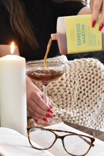 A person pours a brown espresso martini drink from a bottle with a yellow label into a coupe glass. The scene includes a lit white candle, an open book, tortoiseshell eyeglasses, and a cream knit blanket in the background.