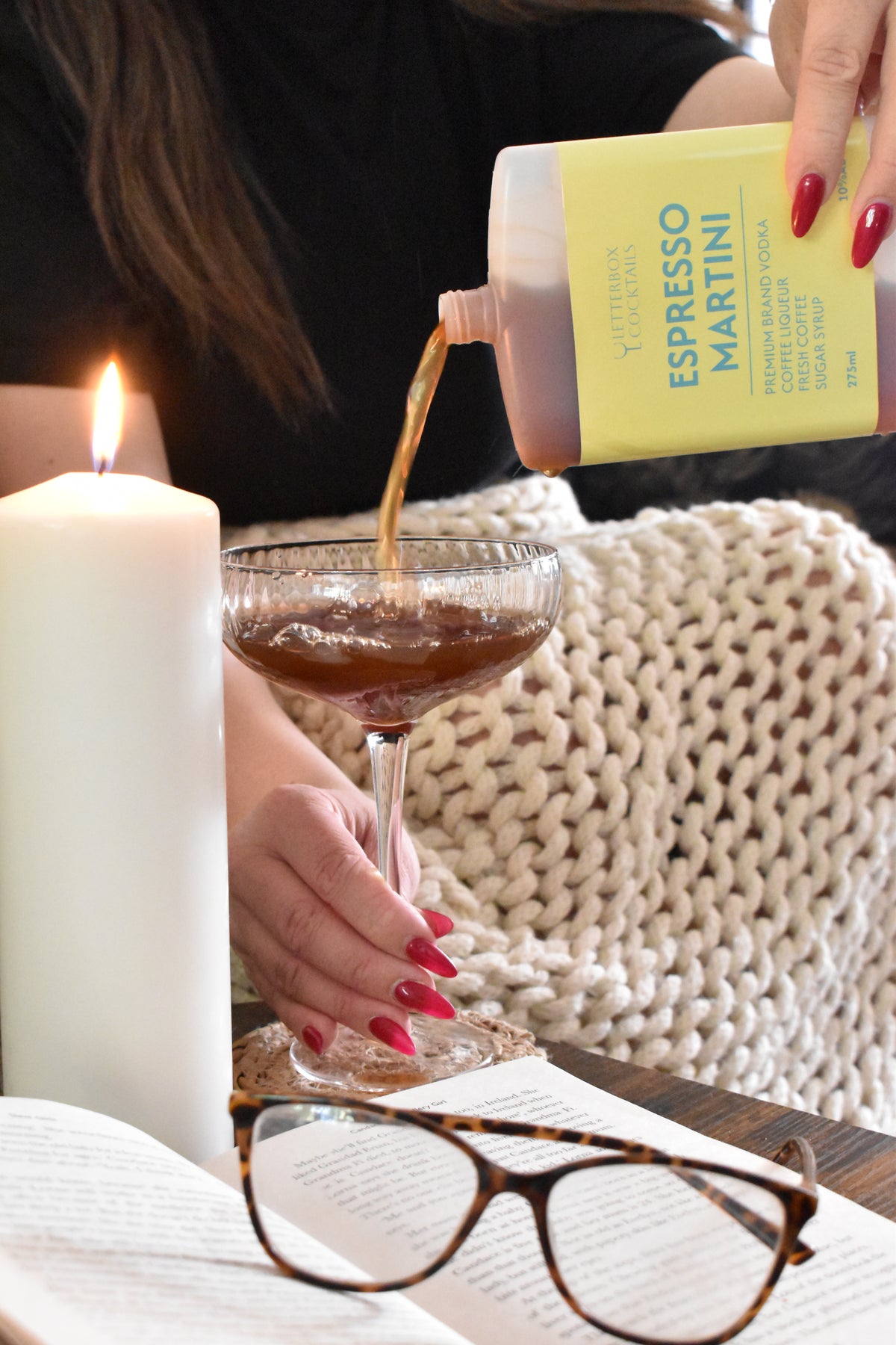 A person pours a brown espresso martini drink from a bottle with a yellow label into a coupe glass. The scene includes a lit white candle, an open book, tortoiseshell eyeglasses, and a cream knit blanket in the background.