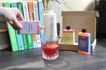 A hand pours a pinkish-red liquid from a bottle labeled 'Fruity Negroni' into a faceted glass. In the background, a cardboard box contains two bottles labeled 'Blood Orange Martini' and 'Italiano Negroni', with colorful books standing behind them on a tabletop.