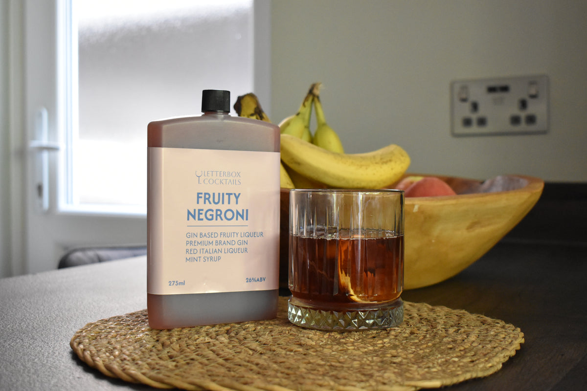 A bottle of Letterbox Cocktails Fruity Negroni gin syrup stands on a woven placemat next to a glass filled with the amber-colored drink, with bananas and a bowl of fruit in the background.