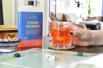 A bottle of Italian Negroni stands on a table next to a hand holding an orange cocktail in a faceted glass, with a blurred living room background and a board game visible in the foreground.
