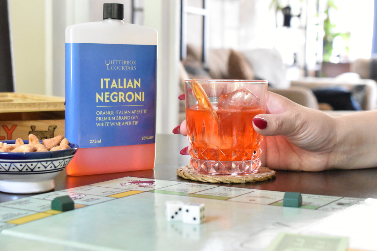 A bottle of Italian Negroni stands on a table next to a hand holding an orange cocktail in a faceted glass, with a blurred living room background and a board game visible in the foreground.