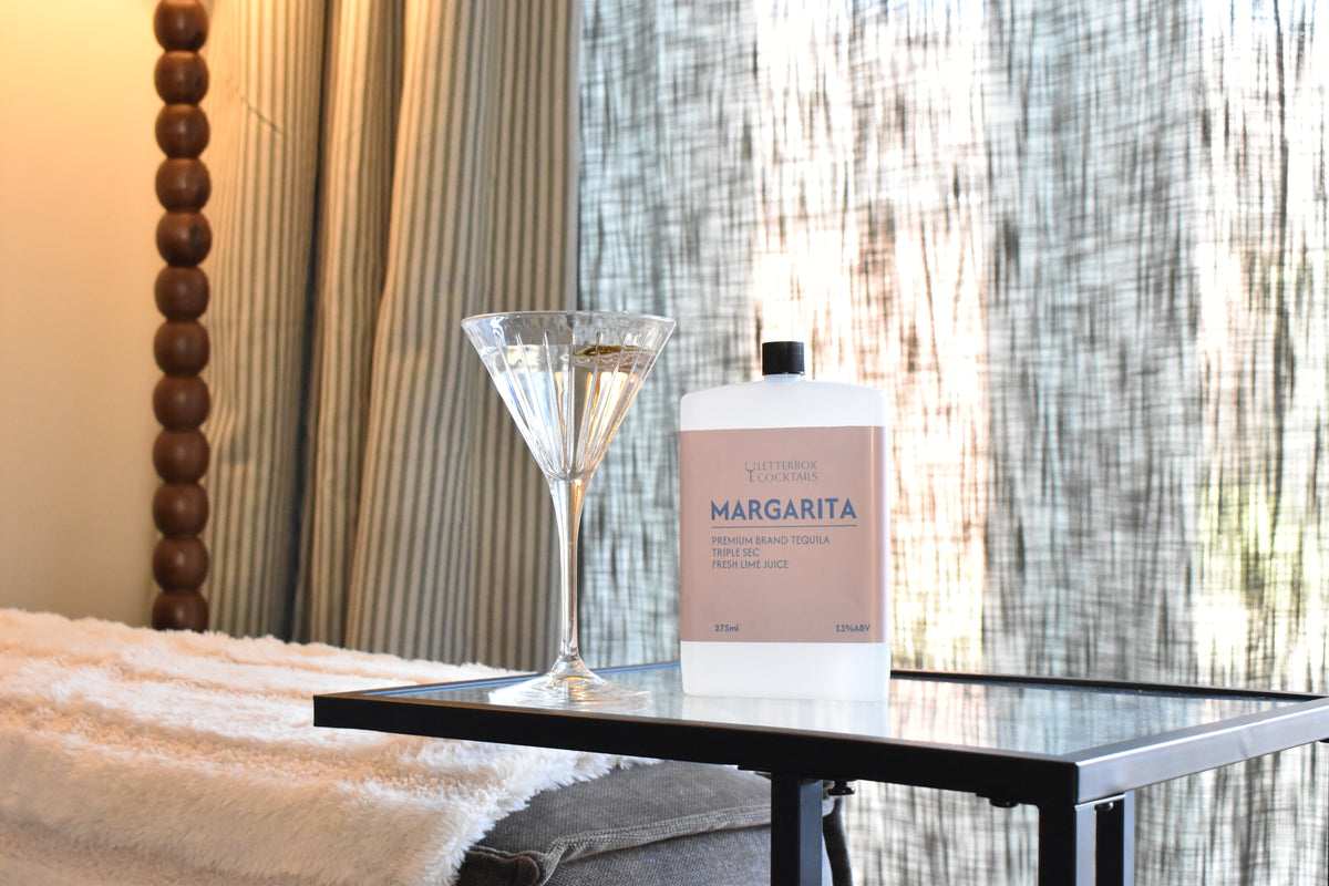 A bottle of Margarita tequila and a martini glass on a black side table in a bedroom setting with curtains and a wooden bed frame visible.