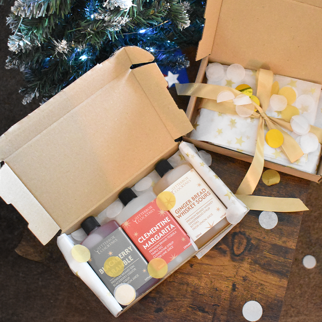 A gift box set on a wooden tabletop, featuring an open box with three mini bottles of letterbox cocktails Christmas flavours  and a closed box wrapped in white paper with gold stars and tied with a gold ribbon.