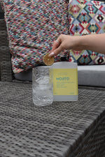 A hand holds a dried orange slice above a clear textured glass and a frosted bottle labeled 'MOJITO' with a pale yellow label, set on a gray woven table with patterned cushions in the background.