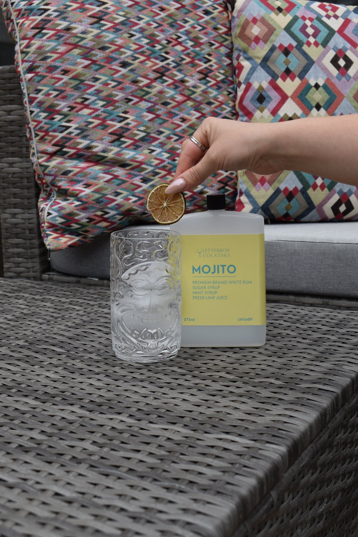 A hand holds a dried orange slice above a clear textured glass and a frosted bottle labeled 'MOJITO' with a pale yellow label, set on a gray woven table with patterned cushions in the background.
