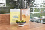 A bottle of Letterbox Cocktails non-alcoholic Passion Daiquiri on a wooden kitchen counter, next to a glass of the fizzy drink and a small bowl of green olives, with a tiled kitchen background.