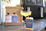 Three bottles of Letterbox Cocktails cocktails displayed in front of their cardboard packaging box on a dark tabletop, with a cocktail glass of yellow liquid and a blurred kitchen appliance in the background.