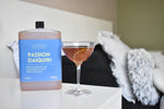 A bottle of Passion Daiquiri rum and passion fruit cocktail stands next to a coupe glass filled with an orange-brown cocktail on a white bedside table, with a blurred bedroom background.