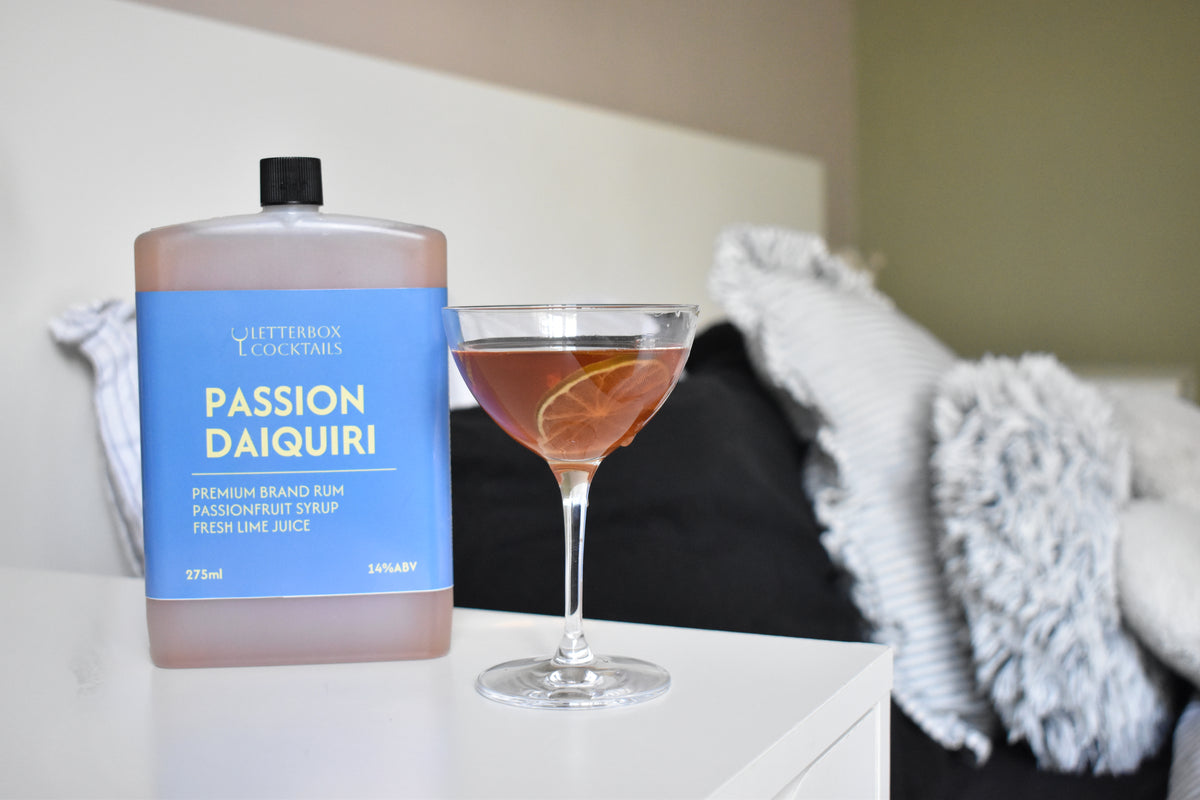 A bottle of Passion Daiquiri rum and passion fruit cocktail stands next to a coupe glass filled with an orange-brown cocktail on a white bedside table, with a blurred bedroom background.