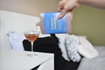 A hand pours a pinkish-orange liquid from a blue bottle labeled 'PASSION DAIQUIRI' into a coupe glass on a white bedside table, with a bed in the background.