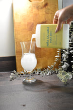 A hand pours a white liquid from a bottle labeled 'PINA COLADA MARTINI' into a clear glass, set against a rustic wooden cutting board and eucalyptus branches.
