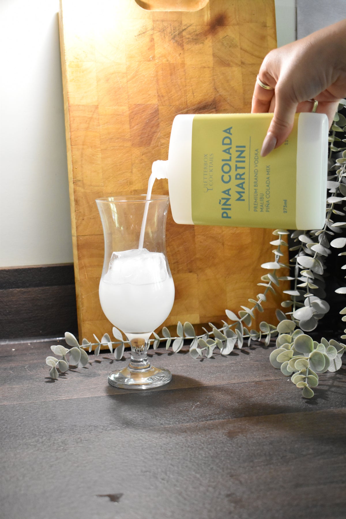 A hand pours a white liquid from a bottle labeled 'PINA COLADA MARTINI' into a clear glass, set against a rustic wooden cutting board and eucalyptus branches.