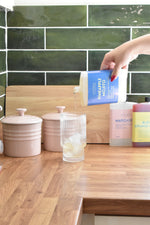 A hand pours a clear liquid from a blue and white bottle labeled PINEAPPLE MOJITO into a ribbed glass on a wooden countertop in a kitchen setting with green subway tiles.