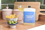 A bottle of Letterbox Cocktails 'Pornstar Martini' premium brand vodka and vanilla syrup fresh lime juice, styled on a wooden countertop with a glass of the yellow drink, olives, and tortilla chips.