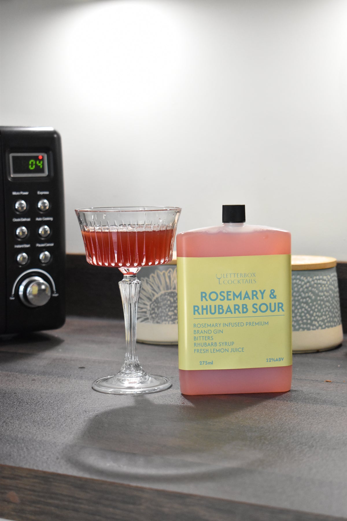 A bottle of Letterbox Cocktails Rosemary & Rhubarb Sour syrup stands on a countertop next to a filled coupe glass with the same pink liquid, with a black appliance and ceramic containers blurred in the background.