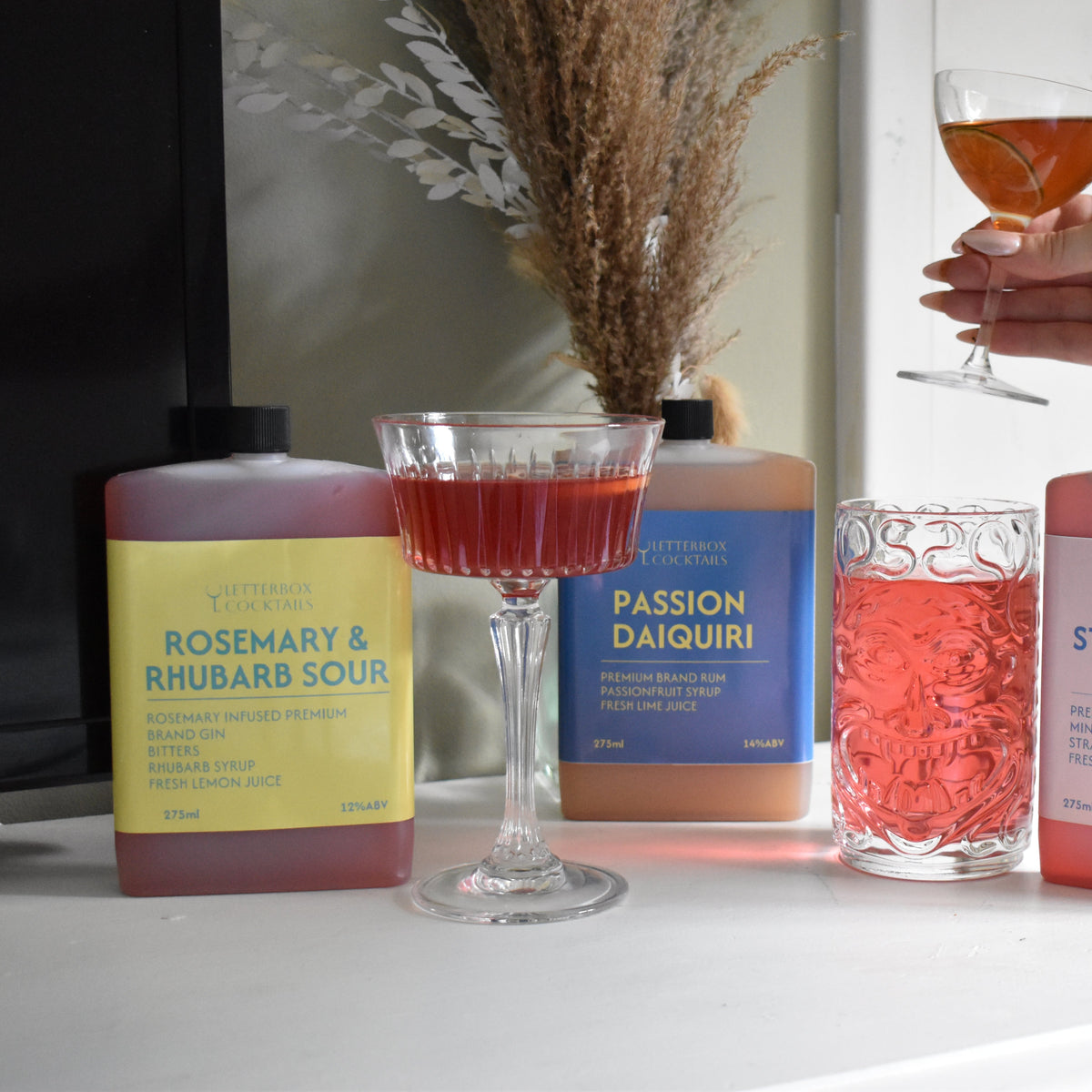 Four bottles of artisanal mixed drinks and three filled glasses on a white table, with a hand holding a glass of the amber drink in the background.