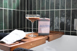 Letterbox Cocktails Salted Caramel Espresso Martini cocktail in a pink bottle, styled on a wooden caddy next to a cocktail coupe glass and a white towel in a bathtub with dark green tiled walls.
