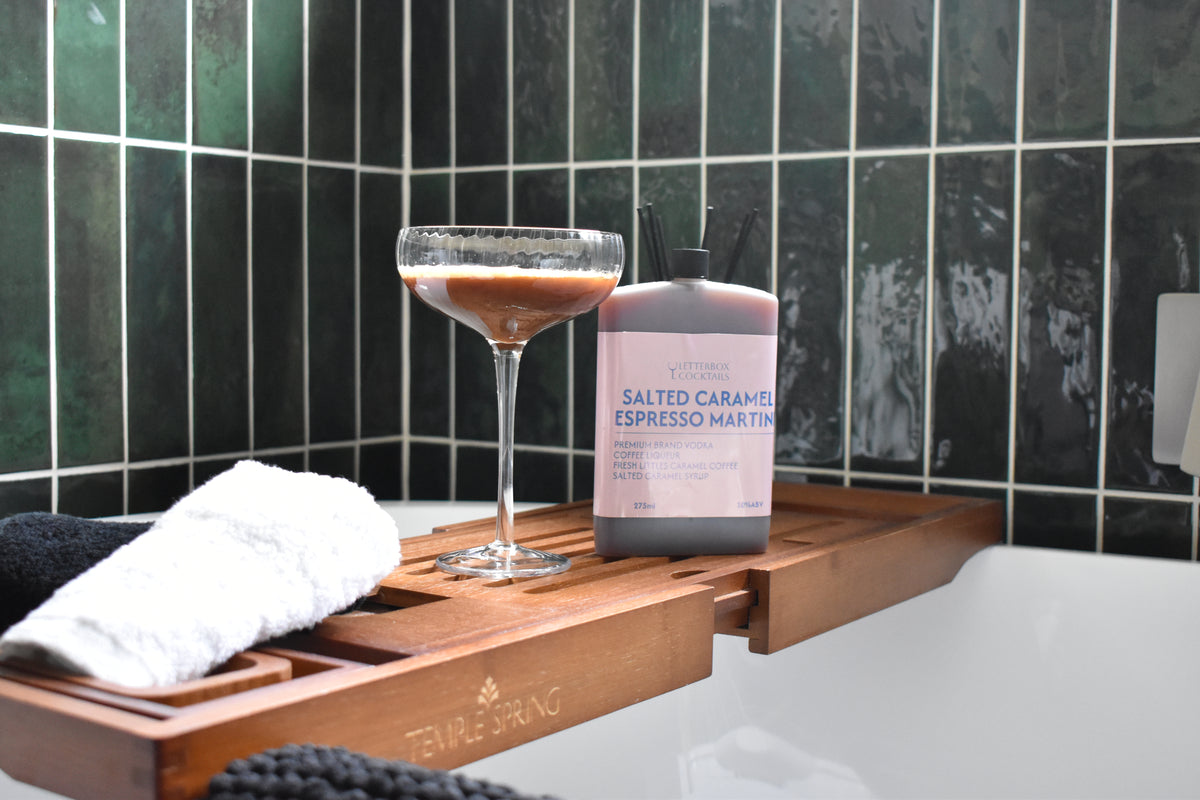 Letterbox Cocktails Salted Caramel Espresso Martini cocktail in a pink bottle, styled on a wooden caddy next to a cocktail coupe glass and a white towel in a bathtub with dark green tiled walls.