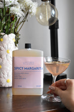 A bottle of Letterbox Cocktails Spicy Margarita mixed drink next to a hand holding a filled coupe glass, set on a dark wooden table with a blurred background of a lamp and white flowers.