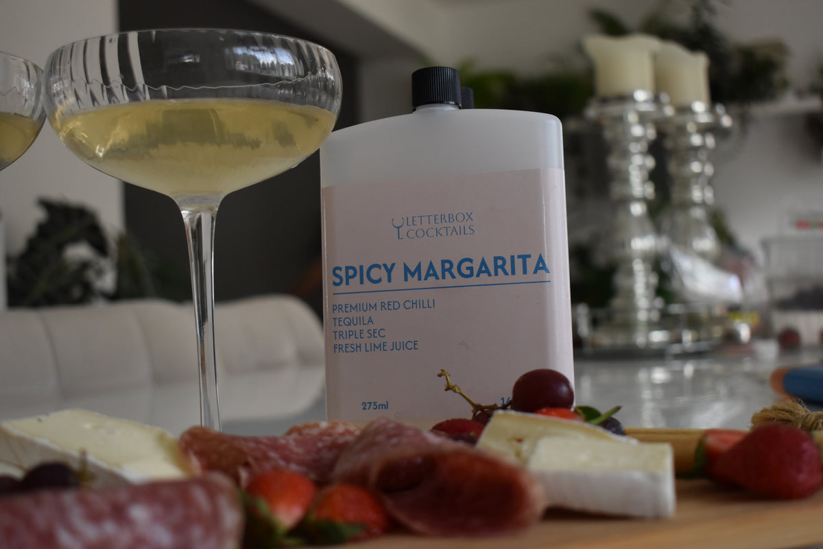 A bottle of Letterbox Cocktails Spicy Margarita cocktail mix in the foreground, with a margarita glass and a charcuterie board in a blurred kitchen background.
