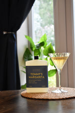 A bottle of Tommy's Margarita cocktail spirit and a filled martini glass sit on a woven placemat on a wooden table, with a window, curtain, and green plant in the background.