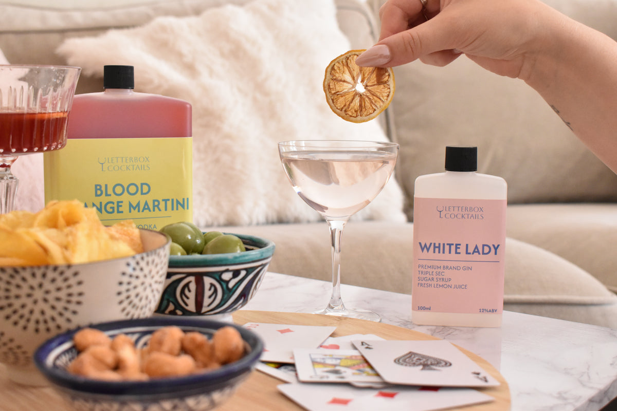 A lifestyle shot of a cocktail setup featuring a bottle of Letterbox Cocktails 'White Lady' (pink) and 'Blood Orange Martini' (yellow) on a marble table, with a hand holding a dried orange slice above a martini glass.