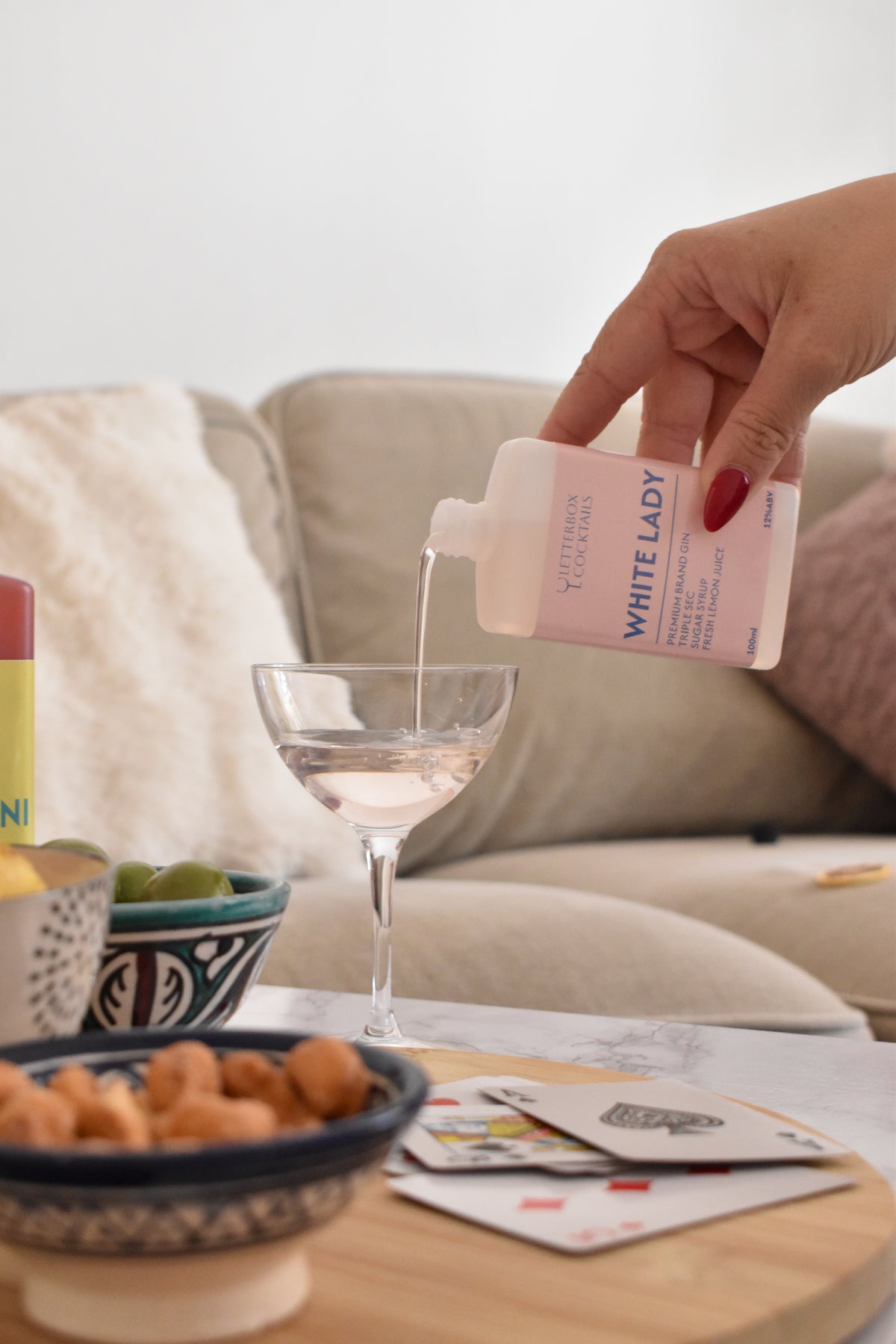A hand with red nail polish pours a clear liquid from a white bottle labeled 'WHITE LADY' into a martini glass, set on a wooden table in a cozy living room environment.