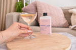 A hand holds a filled coupe glass with a citrus twist next to a bottle of Letterbox Cocktails White Lady on a round wooden tray in a cozy living room setting.