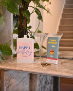A birthday gift set on a marble console table featuring a letterbox cocktail with a pink 'Happy Birthday Emma' label, a clear martini glass, and a chocolate with a blue 'Happy Birthday!' label, set against a blurred background of a staircase and houseplant.