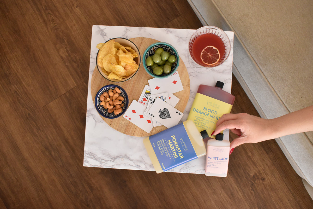 An overhead lifestyle shot of a marble tray styled with snacks, playing cards, and several letterbox cocktails, held by a person's hand on a wooden floor.