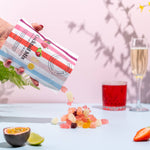 Hand pouring gummy bears from a package labeled 'Gummy Bear Mix' onto a table with a glass of juice and a glass of champagne in the background.