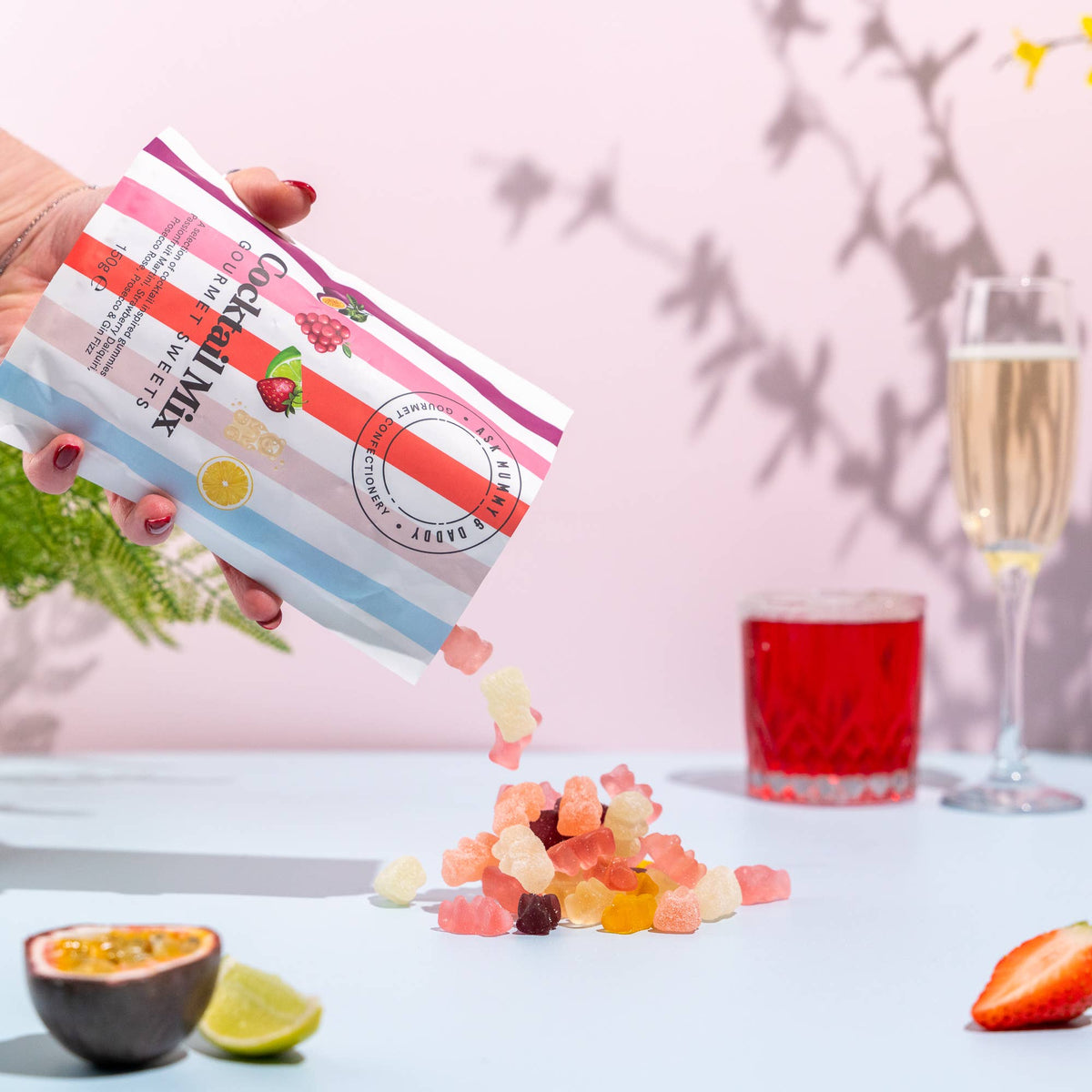 Hand pouring gummy bears from a package labeled 'Gummy Bear Mix' onto a table with a glass of juice and a glass of champagne in the background.