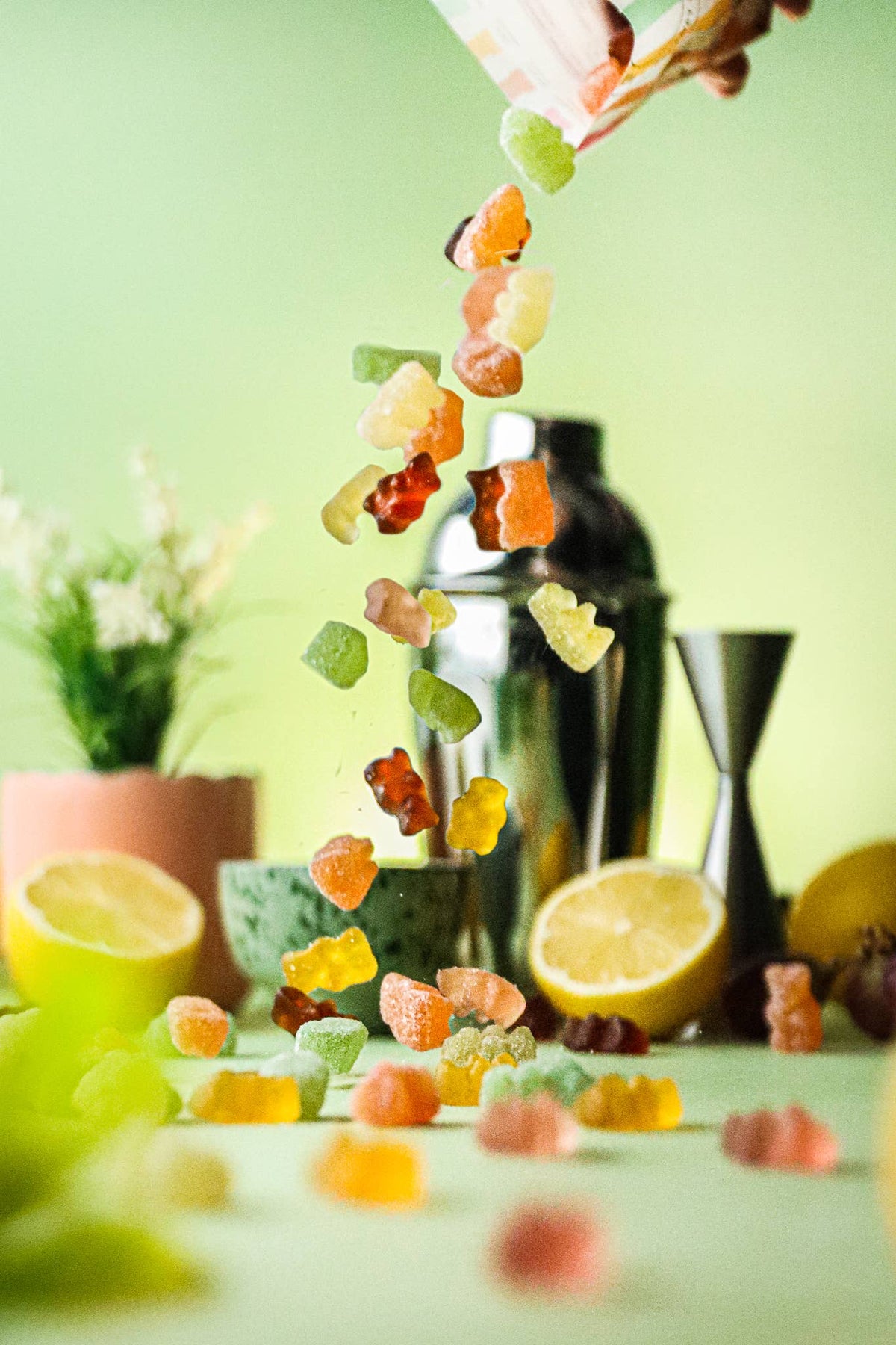 Colorful gummy bears falling from a bottle with a cocktail shaker and lemons on a green background