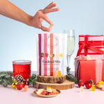 Cocktail mix package with a hand reaching towards it, surrounded by festive items like a glass of red cocktail, champagne flute, and Christmas decorations.