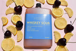 Bottle of whiskey sour with lemons and cherries on a pink background